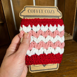 Hand holding a crocheted coffee cozy with red, pink, and white pattern on a wooden surface.